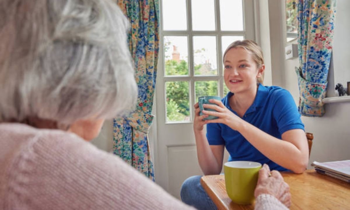 A home health care taker nurse having a cup of tea is sitting in front of an old lady
